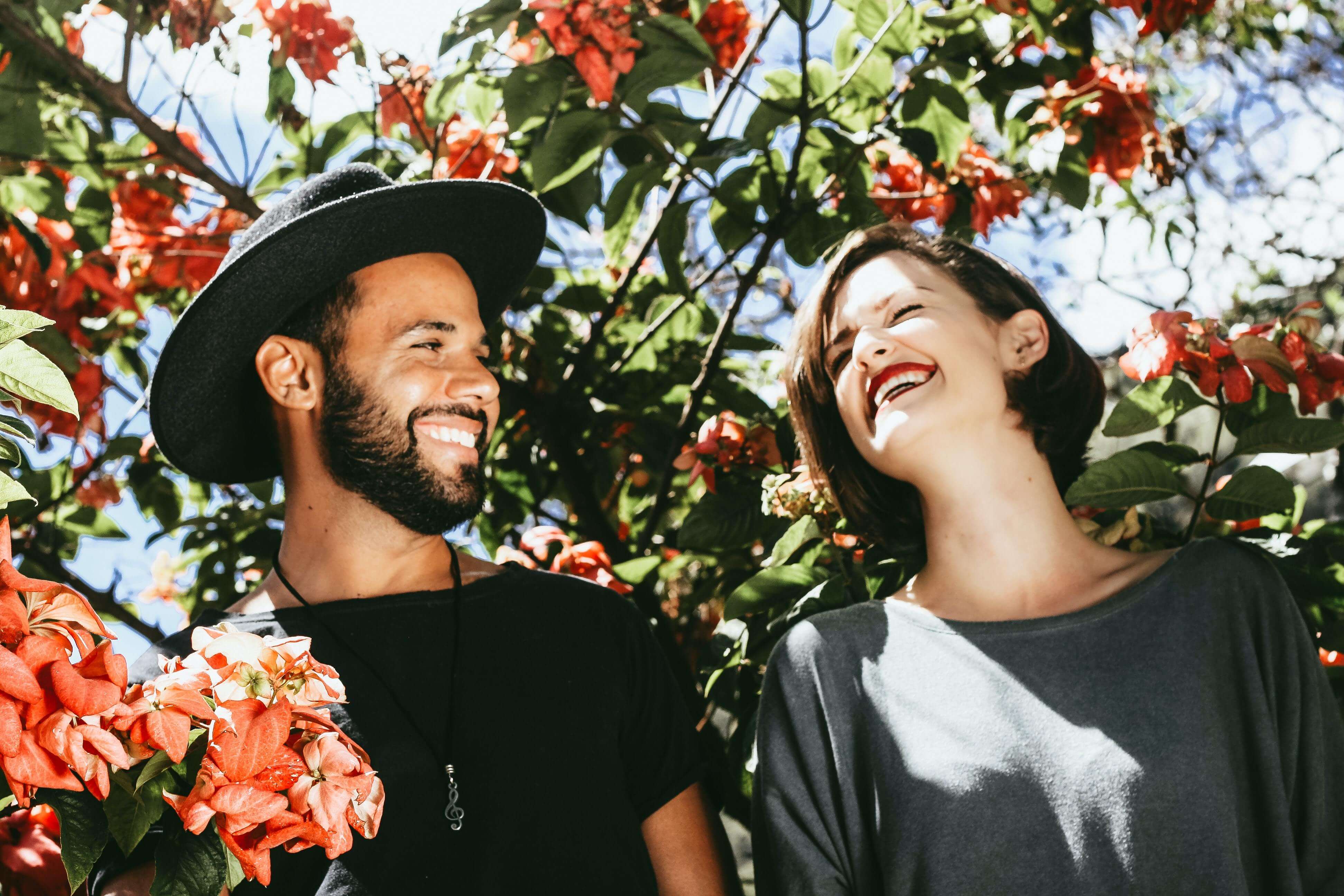 a bearded man in a hat and a woman laughing in front of a tree with pink flowers