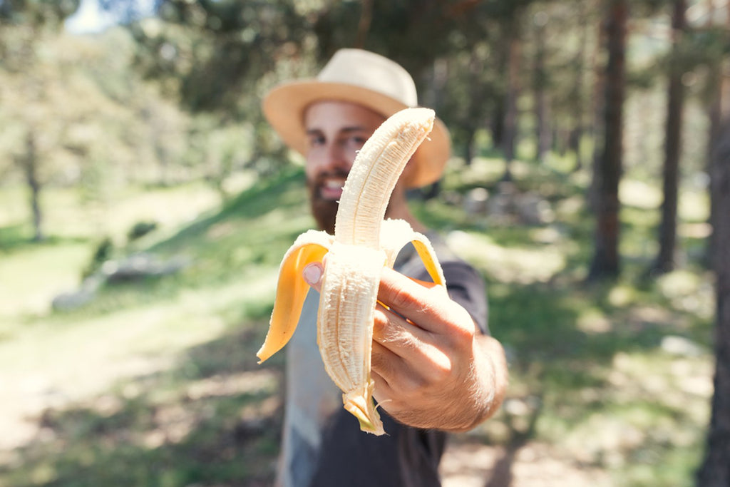 Man smiling wearing a gray shirt and a straw hat is holding forward a peeled banana 