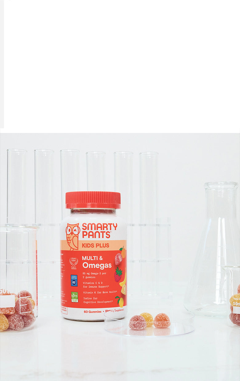 A bottle of Smarty Pants Kids Plus Multi & Omega 3 gummy vitamins stands on a white surface with clear glass beakers and candy pieces around it against a white paneled background.