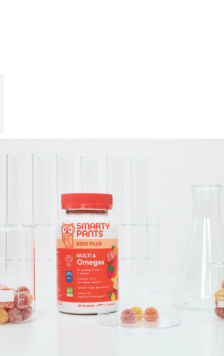 A bottle of Smarty Pants Kids Plus Multi & Omega 3 gummy vitamins stands on a white surface with clear glass beakers and candy pieces around it against a white paneled background.