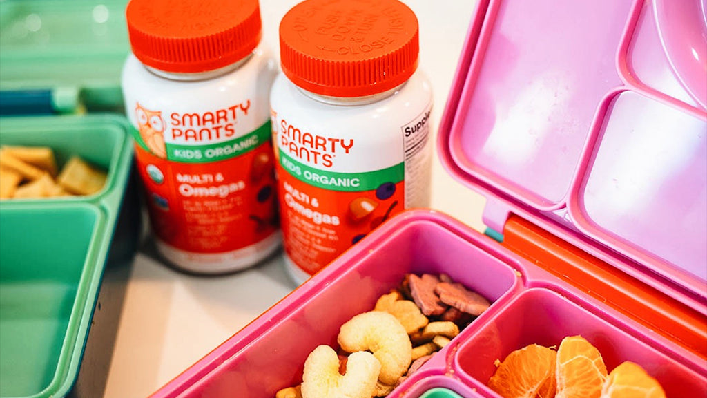 Pink bento box with snacks and two bottles of Smarty Pants vitamins on a wooden surface.